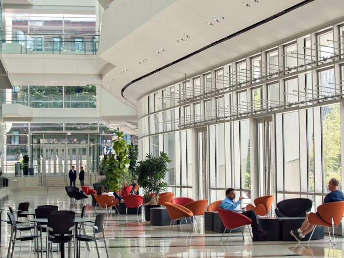 People walking and seated in orange bucket chairs of large modern lobby with curved tall glass walls