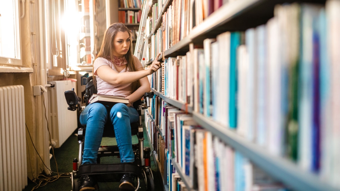 Young woman in a wheelchair selects a book from library shelves