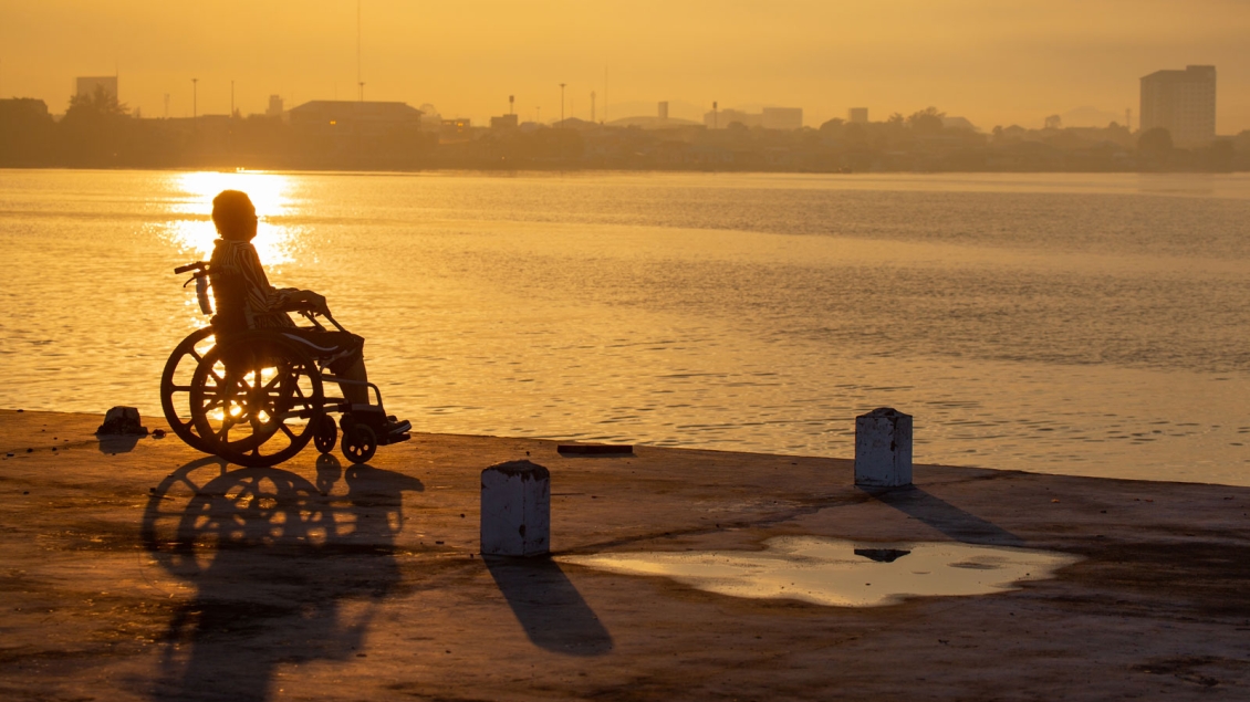 Woman in wheelchair looks out over water & a city skyline at dawn