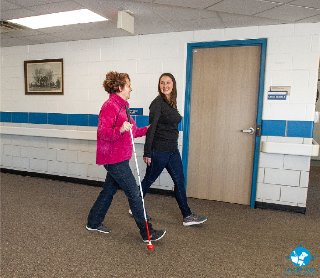 A person who is blind, using a white cane, receiving human guide services in a hallway.